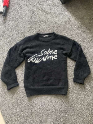 Svart Saint Laurent stickad tröja - Mysig mörkgrå stickad tröja från Saint Laurent med vit, handskriven logga framtill. Tröjan har rund halsringning, långa ärmar och är tillverkad i ett fluffigt, mjukt material som ger en cool och avslappnad vibe.