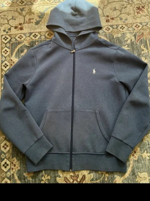 Ralph lauren hoodie - Snygg blå hoodie från Polo Ralph Lauren med dragkedja och klassisk vit broderad logga på bröstet. Hoodien har känguruficka framtill och ribbade muddar vid ärmslut och nederkant. Perfekt för en avslappnad och stilren look. Hör av er vid frågor !