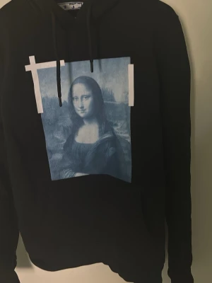 Svart hoodie med Mona Lisa-tryck off white - Svart hoodie med huva och dragsnören. På framsidan finns ett stort blåvitt tryck av Mona Lisa med vita grafiska detaljer. Hoodien har långa ärmar och en klassisk passform. Perfekt för dig som gillar streetwear och unika prints.