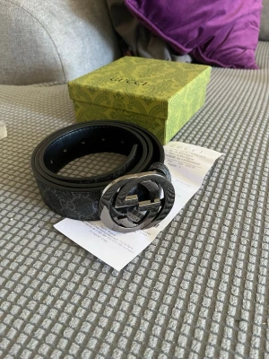 Svart Gucci bälte med GG-spänne - Snyggt svart bälte från Gucci med klassiskt GG-mönster och ett stort, blankt metallspänne i silver. Bältet har en stilren design och levereras med originalask. Perfekt accessoar för att lyfta din outfit.