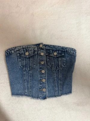 Denim tubtopp med knappar - Snygg tubtopp i denim med knappar framtill och två fejkfickor. Toppen har fransig nederkant och smock baktill för en bekväm passform. Perfekt för en trendig och avslappnad look.