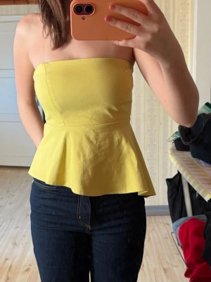 Gul axelbandslös peplum-topp - Snygg gul axelbandslös topp med peplumskärning. Perfekt för dig som vill ha en färgglad och trendig look. Toppen är figurnära upptill och har en utsvängd nederdel som ger en fin siluett.