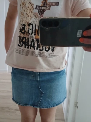 Blå jeanskjol i kort modell - Snygg blå jeanskjol i klassisk kort modell med femficksdesign och knapp- samt dragkedjestängning framtill. Midjemått 37 cm rakt över. Har ett håll inne men syns inte när man har den på