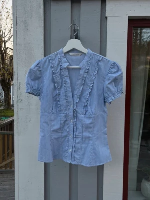 Blårandig blus med volang från Zara - Söt blårandig blus från Zara med korta puffärmar och volangdetaljer längs knäppningen. Blusen har en v-ringad hals och tunna vita ränder på blå botten. Perfekt för en trendig och avslappnad look.