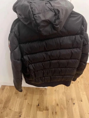 Svart quiltad pufferjacka med luva - Snygg svart pufferjacka med luva och quiltad design. Jackan har en patch på ärmen och breda kanaler för extra värme. Perfekt för kalla dagar och har en modern, oversized passform.