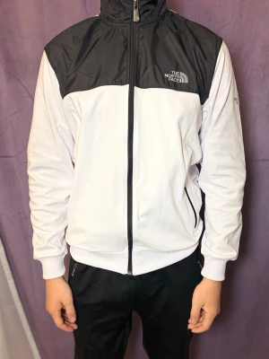 Vit och svart vindjacka The North Face - Snygg vindjacka från The North Face i vitt med svart axelparti och detaljer. Jackan har hel dragkedja, två sidofickor med dragkedja och hög krage. Tillverkad i lätt material som passar perfekt för blåsiga dagar. Logga på bröstet och ärmen.