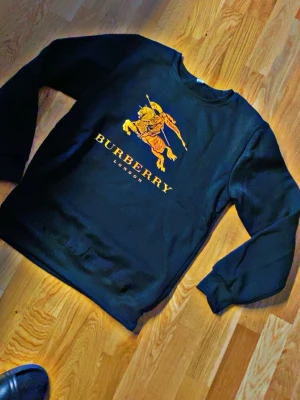 Svart Burberry tröja  ny at m passar s  - Svart sweatshirt från Burberry med stort gult och orange tryck av en riddare på häst och texten 'BURBERRY LONDON' på bröstet. Tröjan har rund hals och långa ärmar, perfekt för en stilren och trendig look. Ny liten i storlek passar 165/175