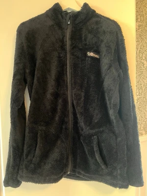 Svart teddyfleece från Ulmax - Mysig svart teddytröja från Ulmax med dragkedja framtill och två fickor, mjukt och fluffigt material. Perfekt för kyliga dagar när du vill vara både varm och stilren.