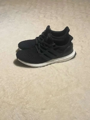 Svarta Adidas Ultraboost sneakers - Svarta Adidas Ultraboost sneakers med stickad ovandel och vita Boost-sulor. Skorna har klassiska tre ränder på sidan och en sportig, låg profil. Perfekta för dig som gillar streetstyle och komfort. Ovandelen är i mesh för extra andningsförmåga. Har storlekar 42,43 och 44 