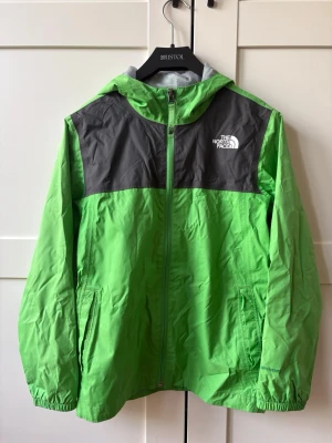 Grön vindjacka från The North Face (barnstl M) - Snygg grön vindjacka från The North Face med svarta axelpartier och vit logga på bröstet. Jackan har huva, dragkedja framtill och två sidofickor. Tillverkad i lätt och slitstarkt material med meshfoder för extra komfort. Perfekt för blåsiga dagar.