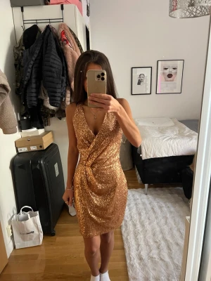 Glittrig guldig omlottklänning - Snygg kort glittrig klänning i guld med omlott-design och djup v-ringning. Klänningen är ärmlös och har en figurnära passform, perfekt för att sticka ut. Materialet är täckt av paljetter som ger en lyxig känsla.