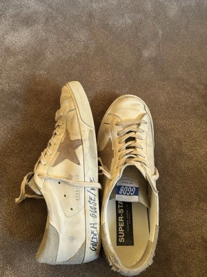 Beige Golden Goose Superstar sneakers - Säljer ett par beige Golden Goose Superstar sneakers med den ikoniska stjärnan på sidan och textdetaljer på hälen. Skorna har vita snören, gummisula och är tillverkade i läder med slitna detaljer för en cool vintage-look.
