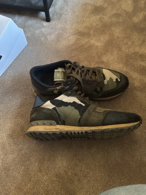 Valentino camo sneakers i grönt och svart - Snygga sneakers från Valentino med camouflage-mönster i grönt, svart och beige. Skorna har snörning, gummisula och detaljer i mocka och textil. Perfekta för dig som vill sticka ut med en cool streetstyle-look.