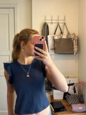 Mörkblå topp med volangärmar Lindex - Supersöt mörkblå topp från Lindex med volangdetaljer på ärmarna. Toppen är lätt croppad och har en strukturerad yta som ger en cool känsla. Perfekt att styla med jeans eller kjol för en trendig look.
