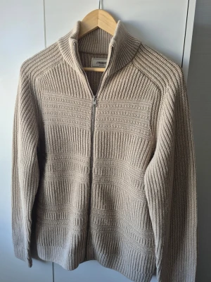 Beige stickad ziptröja - Säljer en beige stickad tröja med dragkedja framtill och hög krage. Tröjan har ett snyggt ribbat mönster och är tillverkad i ett tjockare material som passar perfekt när det är kyligt ute. Långärmad och riktigt skön att ha på sig.