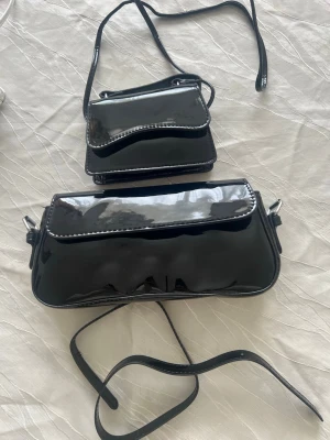 Svarta lackade handväskor med axelrem - Två stilrena svarta handväskor i glansigt lackmaterial med justerbara axelremmar. Väskorna har enkel design med lock och silvriga detaljer på sidorna. Perfekta för att lyfta en outfit med en modern touch. Pris kan diskuteras 