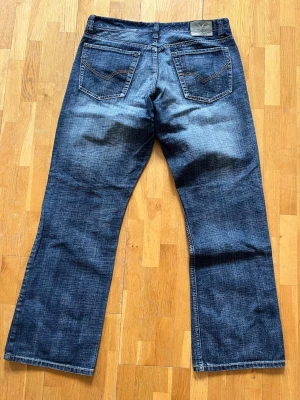 Blå bootcut jeans från River Island - Säljer ett par klassiska blå bootcut jeans från River Island med snygga slitningar och kontrastsömmar. Jeansen har fem fickor och en avslappnad passform med bredare ben. Perfekta för en cool och avslappnad stil.