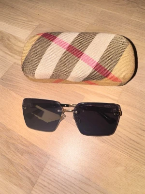 Burberry fyrkantiga solglasögon - Snygga solglasögon från Burberry med fyrkantiga svarta glas och guldfärgade detaljer. Kommer med klassiskt fodral i Burberrys rutiga mönster och originalkartong. Perfekt accessoar för att lyfta din stil. Köpt från OUNASS Kuwait.
