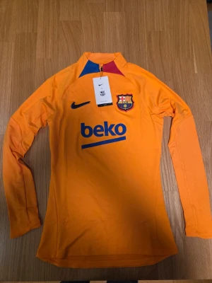 Träningströja Nike FC Barcelona Strike Women s Soccer Drill Top storlek XS - Träningströja från FC Barcelona i orange med långa ärmar och dragkedja vid halsen. Tröjan har klubbens emblem på bröstet tillsammans med Nike-logotypen. På ryggen finns texten "beko" i blått. Ärmarna har ett diskret mönster. Säsong 2021/22