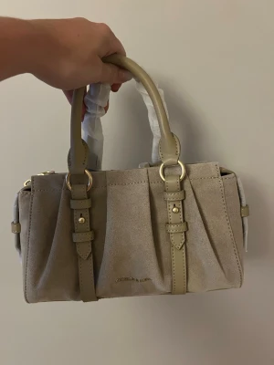 Beige handväska Charles & Keith - Stilren beige handväska från Charles & Keith i mockaliknande material med detaljer i ljusbeige läder och guldfärgade metalldetaljer. Väskan har två handtag och dekorativa spännen framtill. Perfekt för dig som gillar en klassisk och trendig look.