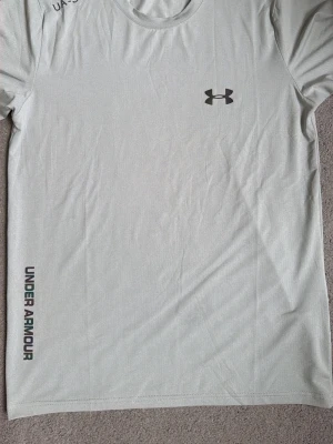 Ljusgrå träningströja Under Armour - ((((Oanvänd)))Snygg ljusgrå träningströja från Under Armour med logga på bröstet och märkesnamn tryckt längs sidan. Tillverkad i ett lätt och snabbtorkande funktionsmaterial, perfekt för gym eller löpning.