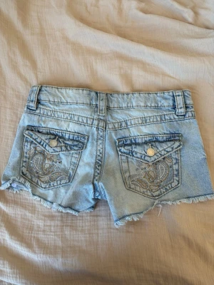 Ljusblå jeansshorts med broderi - Snygga ljusblå jeansshorts från Gina Tricot med slitna kanter och broderade detaljer på bakfickorna. Klassisk femficksmodell med knapp och dragkedja framtill. Perfekta för varma dagar och en avslappnad stil.