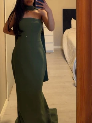 Mörkgrön axelbandslös långklänning - Säljer en elegant mörkgrön långklänning med axelbandslös design och matchande halsbandsscarf. Klänningen har en figurnära passform. Perfekt för dig som vill sticka ut med en stilren look.