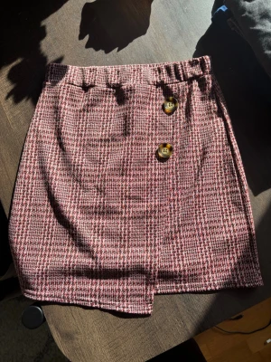 Rutig kortkjol med knappar - Säljer en snygg kortkjol med rutigt mönster i rosa, svart och vitt. Kjolen har två dekorativa knappar framtill och en liten slits. Perfekt för dig som gillar trendiga och klassiska plagg med coola detaljer.