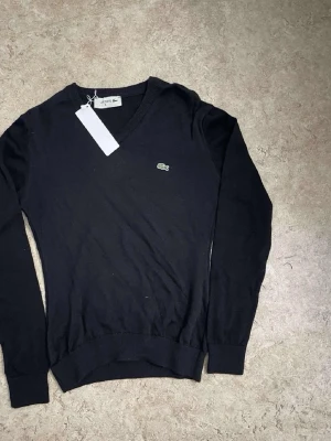Svart v-ringad tröja från Lacoste - Snygg svart v-ringad tröja från Lacoste med det klassiska krokodilmärket broderat på bröstet. Tröjan har långa ärmar och ribbade muddar vid ärmslut och nederkant. Perfekt för dig som gillar stilrena plagg med ikonisk logga.