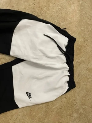 Nike svartvita mjukisbyxor - Snygga mjukisbyxor från Nike i svart och vitt med logga på benet. Byxorna har elastisk midja med dragsko och är tillverkade i mjukt material som passar perfekt för chill och sport. Färgblock-designen ger en modern look.