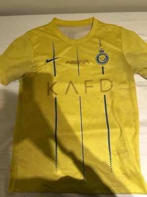 Gul Al Nassr matchtröja Nike - Snygg gul matchtröja från Al Nassr med blå detaljer och klubbmärke på bröstet. Tröjan har korta ärmar, rund hals och är tillverkad i ett lätt, ventilerande material perfekt för fotboll eller träning. Stora tryck på framsidan och vertikala blå linjer ger en sportig look.