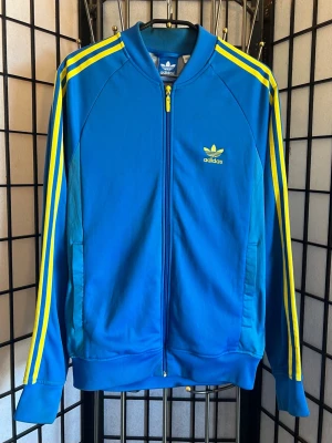 Adidas zip track jacket blå/gul 🇸🇪 Y2K vibe – perfekt till sommaren - Sjuk adidas track jacket i klar blå med gula stripes 💙💛 Riktig Sverige-feel – perfekt inför sommaren, festivaler & fotboll ⚽🇸🇪  Clean look med trefoil logo på bröstet + klassiska stripes längs ärmarna. Funkar lika bra oversized som mer fitted beroende på stil.  Y2K/retro sport vibe som är 🔥 just nu – easy outfit piece.  Storlek: M (Fits M, kan funka S oversized)  Skick: Bra skick – lite slitage på zipper pull (se bild) men inget som stör.  💙 Perfekt till: – VM/sommar vibes 🇸🇪 – streetwear fits