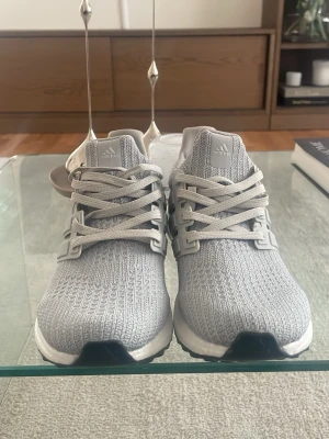 Adidas UltraBoost grå sneakers - Säljer ett par Adidas UltraBoost sneakers i ljusgrå färg med stickad mesh-överdel och klassiska tre ränder på sidan. Skorna har en vit Boost-sula och grå detaljer på häl och snörning. Perfekta för dig som vill ha både stil och komfort.
