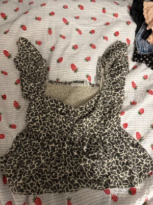 Leopardmönstrad blus med volang - Säljer en söt blus med leopardmönster i svart och beige. Blusen har volangdetaljer på axlarna och en smockad rygg för extra komfort. Materialet är en mix av rayon, bomull och linne. Perfekt för dig som vill ha en trendig och luftig topp.