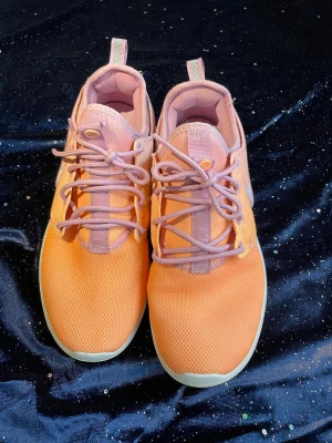 Nike sneakers i orange och lila - Snygga Nike sneakers med meshmaterial i orange och lila nyanser. Skorna har lila snörning, vit sula och klassisk Nike-logga på sidan. Lågt skaft och bekväm passform, perfekta för dig som gillar färgstarka och sportiga skor.