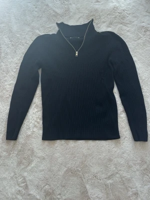 Svart ribbstickad tröja med dragkedja - Säljer en svart ribbstickad tröja med hög krage och dragkedja framtill. Tröjan har långa ärmar och är tillverkad i ett mjukt stickat material som ger en snygg och bekväm passform. Perfekt för att skapa en cool och avslappnad look.