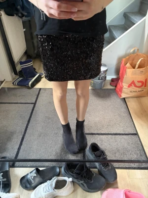 Svart paljettkjol med elastisk midja - Säljer en svart kortkjol täckt med glittriga paljetter. Kjolen har en rak passform och elastisk midja som gör den bekväm att bära. Perfekt för dig som vill sticka ut med en glammig look.