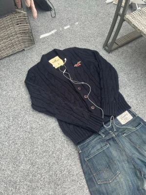 Hollister cable knit cardigan XL old money - Mörkblå Hollister cable knit cardigan i XL. Grovstickad knappkofta med broderad logo och riktigt fin preppy/old money vibe. Ullblandning så den har tung, kvalitativ känsla och är perfekt för höst/vinter. Funkar snyggt med jeans, chinos eller layered över skjorta. Bra skick och riktigt clean vintage piece.