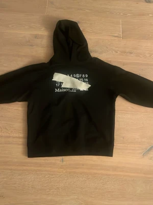 Svart hoodie Maison Margiela - Svart hoodie från Maison Margiela med tryck framtill, samt ett distinkt tejp-liknande motiv över loggan. Klassisk huva och lång ärm. Hoodien har en avslappnad passform och är tillverkad i mjukt bomullsmaterial. Hör av er vid intresse