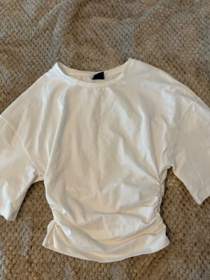 Vit t-shirt med snörning i sidan - Säljer en vit t-shirt med korta ärmar och cool snörningsdetalj i sidan som ger en snygg twist. Perfekt för dig som vill ha något enkelt men ändå lite extra. Materialet är mjukt och känns som bomull.