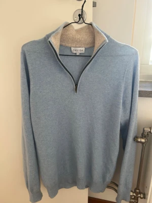 Kashmir halvzip tröja från Davida - Ljusblå långärmad tröja från Davida med halvzip och hög krage. Tröjan är tillverkad i kashmir   och har en kontrastfärgad insida på kragen i beige. Perfekt för en chill och stilren look.