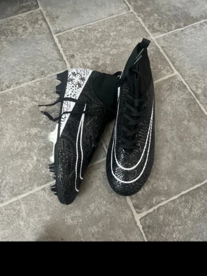 Nike Mercurial fotbollsskor svart/vit - Säljer ett par Nike Mercurial fotbollsskor i svart med vita detaljer och mönster. Skorna har hög krage, snörning och en mönstrad ovandel i syntetmaterial. Sulan har dobbar för bra grepp på planen och coola grafiska inslag på hälen.