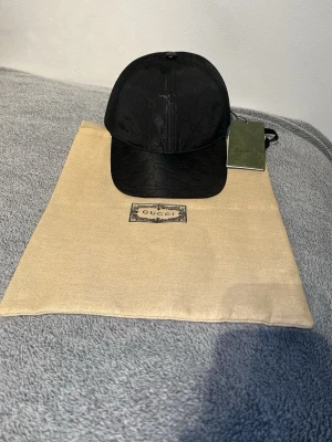 Svart keps från Gucci med monogram - Snygg svart keps från Gucci med klassiskt GG-monogram i ton-i-ton. Kepsen har böjd skärm, mesh baktill och justerbar rem. Tillverkad i en mix av textil och mesh för en modern look. Perfekt accessoar för dig som gillar exklusiv streetwear.