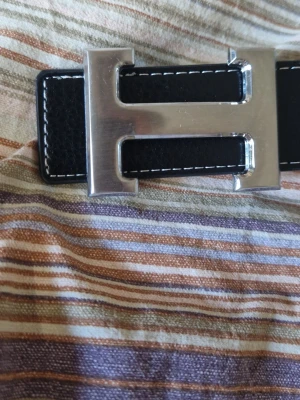 Svart skärp med H-spänne - (Helt nytt och oanvänt) Snyggt svart bälte med ett stort, silvrigt H-format spänne och vita sömmar längs kanterna. Perfekt accessoar för att lyfta vilken outfit som helst. Klassisk och stilren design.