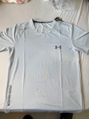 Ljusgrå träningströja Under Armour - Storlek xl men är liten i storlek. Snygg ljusgrå träningströja från Under Armour med korta ärmar och rund hals. Tröjan har logotyp på bröstet och tryck på ärm och nederkant. Tillverkad i ett lätt och snabbtorkande funktionsmaterial som passar perfekt till träning.