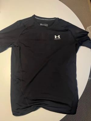 Svart Under Armour HeatGear tröja - Säljer en svart kompressionströja från Under Armour HeatGear. Tröjan har korta ärmar, rund hals och logotyp på bröstet. Perfekt för träning och sport, tillverkad i ett lätt och stretchigt material som andas. (Reminder) tröjan sitter tight som är perfekt ifall man gillar flexa bicepsen på gymmet.