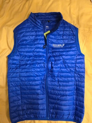 Blå dunväst från Gul & Blå - Snygg blå dunväst från Gul & Blå med quiltad design och dragkedja framtill. Västen har hög krage och logotyp på bröstet. Perfekt för lager-på-lager och håller dig varm under kyliga dagar. Materialet är lätt och smidigt.