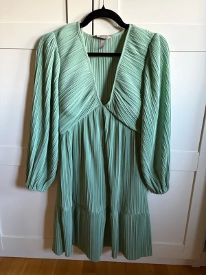 Ljusgrön plisserad klänning H&M - Trendig ljusgrön klänning från H&M med plisserat tyg och v-ringning. Klänningen har långärmade puffärmar och en lös passform som ger en avslappnad men stilren look. Perfekt för vår och sommar när du vill sticka ut lite extra.