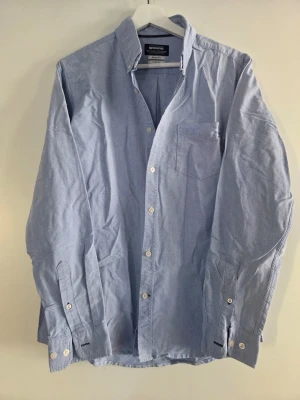 Ljusblå skjorta från Oscar Jacobson - Klassisk ljusblå skjorta från Oscar Jacobson i bomull. Skjortan har lång ärm, bröstficka och vita knappar framtill. Perfekt till både jeans och chinos för en clean och stilren look.