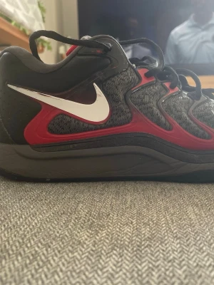 Kevin durant svart/röd - Säljer ett par snygga Kd’s med svart och grå mesh, röda detaljer och vit swoosh på sidan. Skorna har en sportig design med kraftig sula och snörning. Perfekta för dig som vill ha ett par snygga basketskor och komfort till träningen eller basketmatcher.